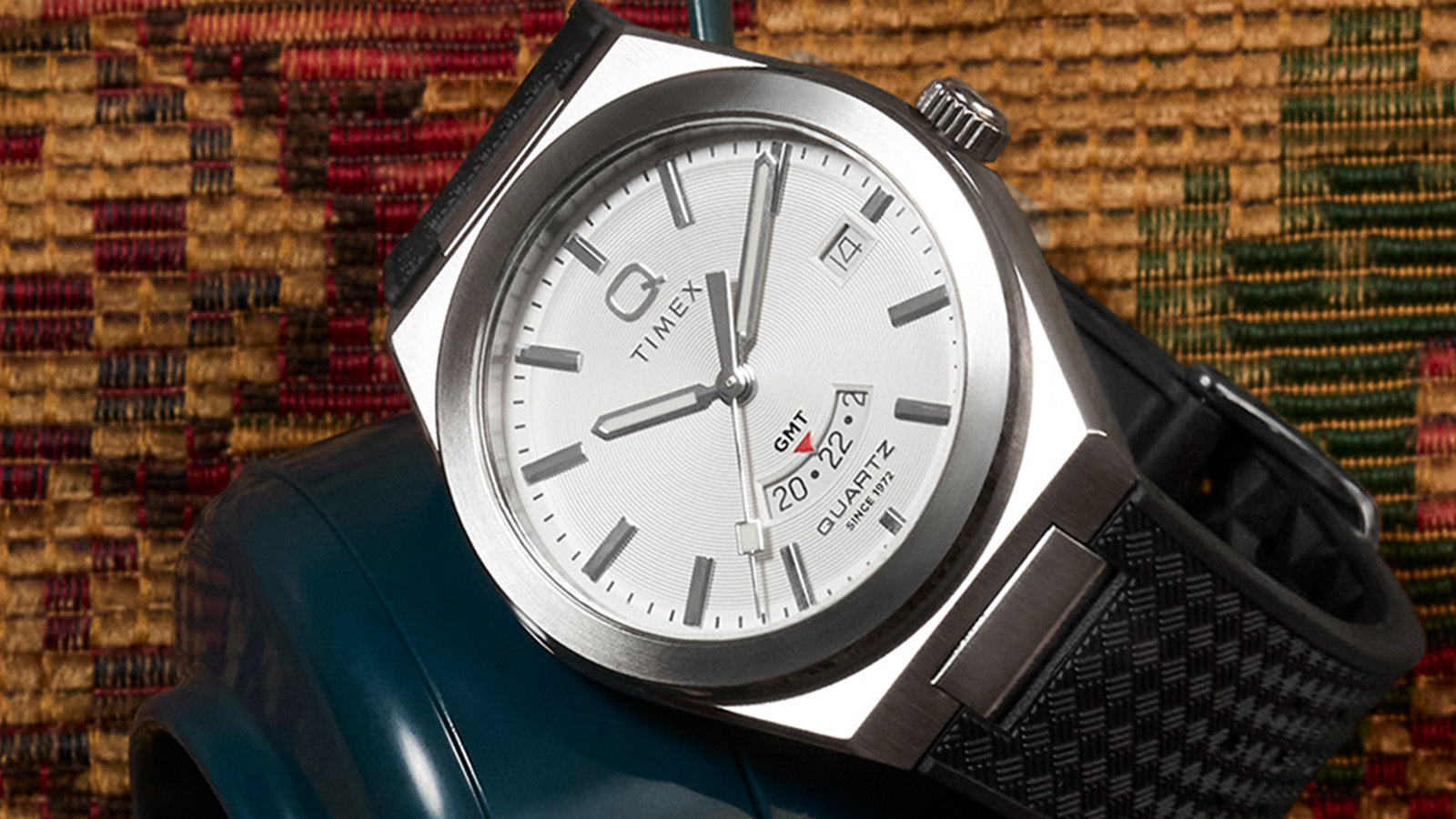 Profile view of the Q Timex® Continental GMT showing its brushed stainless-steel case, slim profile, and engraved “Q” logo crown with black strap attachment.