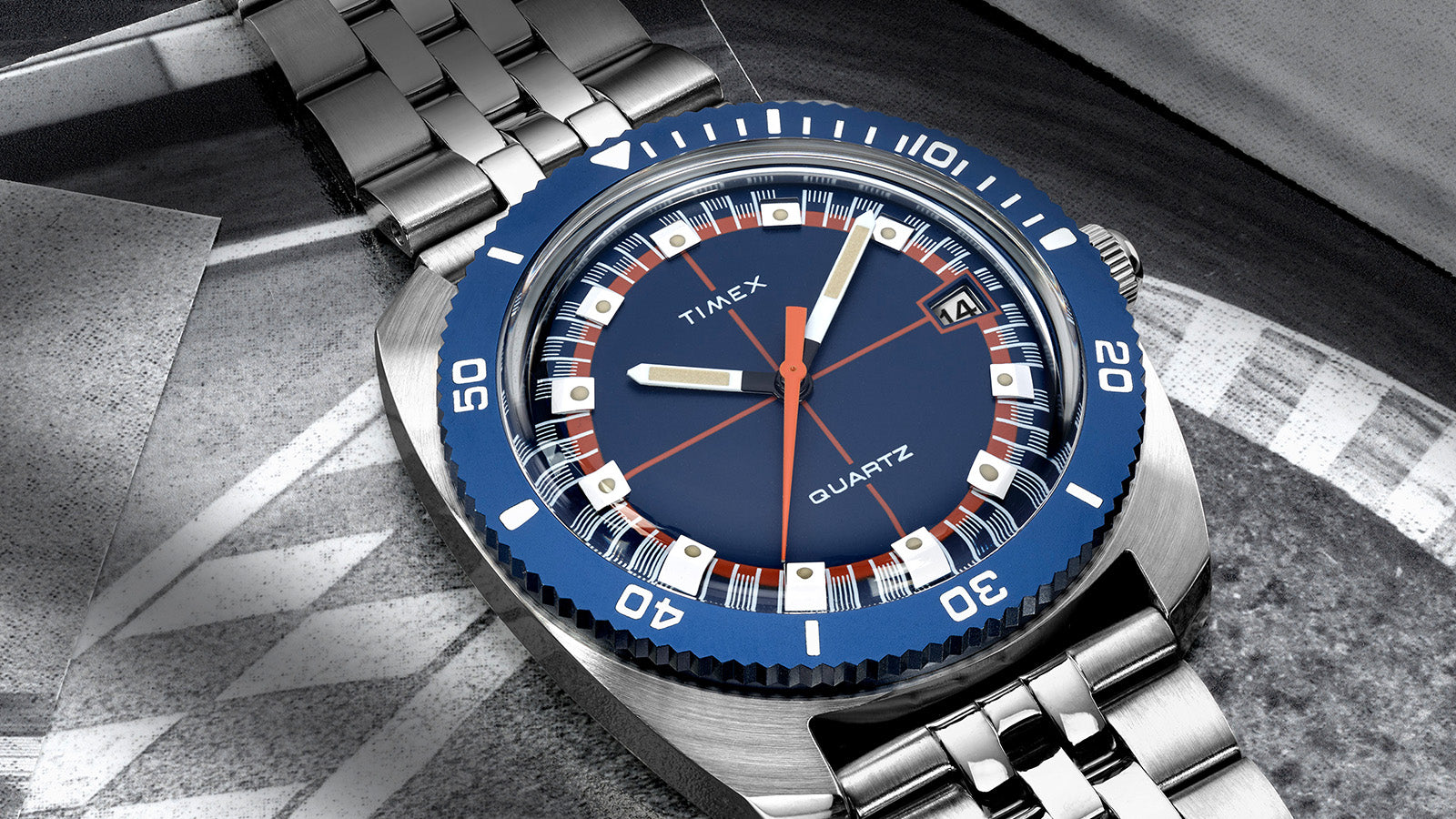 Detailed macro of the Q Timex® 1971 Velocity watch face, featuring a navy blue dial, orange crosshair design, and silver hour markers under a domed crystal.