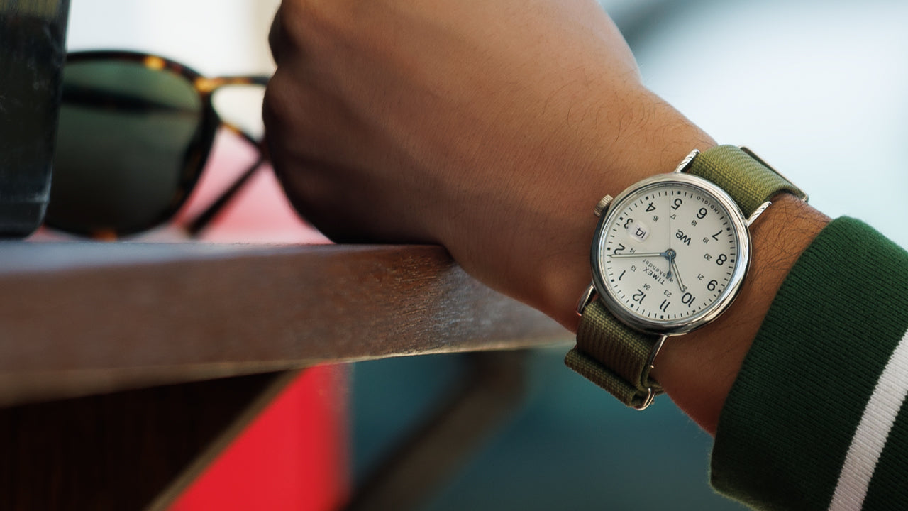 Timex Weekender watch with green strap worn on a wrist, styled casually with a green and white striped sweatshirt near sunglasses on a table.