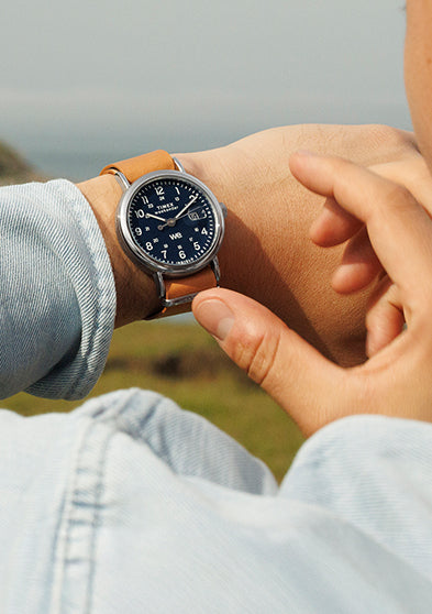 Lifestyle shot of a Timex Weekender with blue dial and brown leather strap, worn on wrist while adjusting time outdoors with a lighthouse in the background.