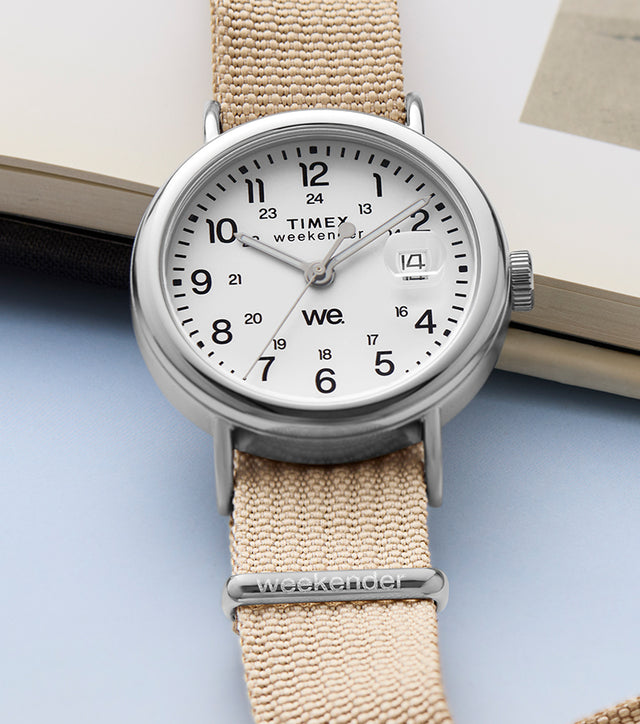 Timex Weekender watch with a beige fabric strap and white dial, placed beside an open book showing a vintage beach photograph.