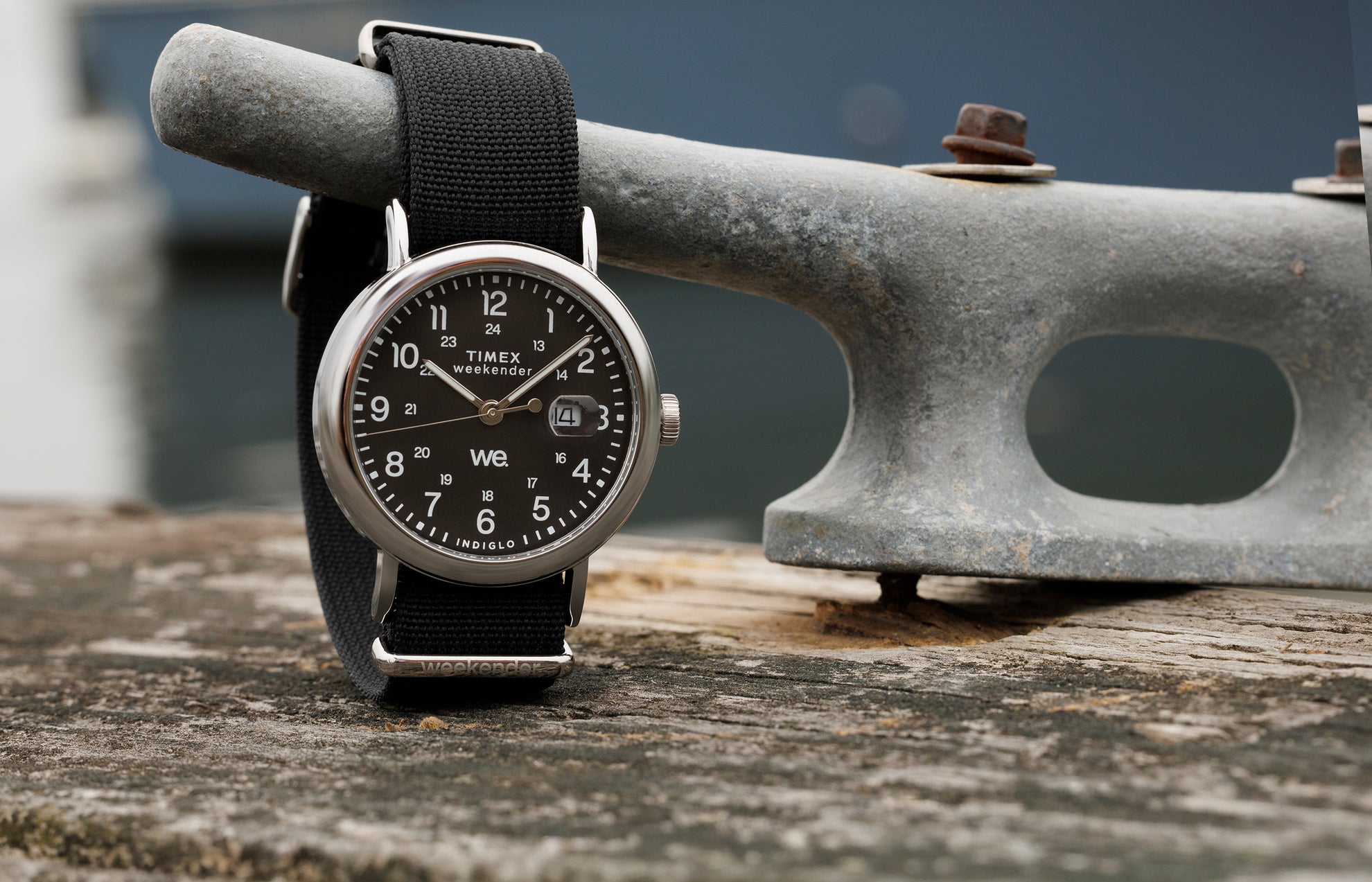Timex Weekender watch with black dial, silver-tone case, and black fabric strap resting on a weathered wooden dock beside a metal cleat.