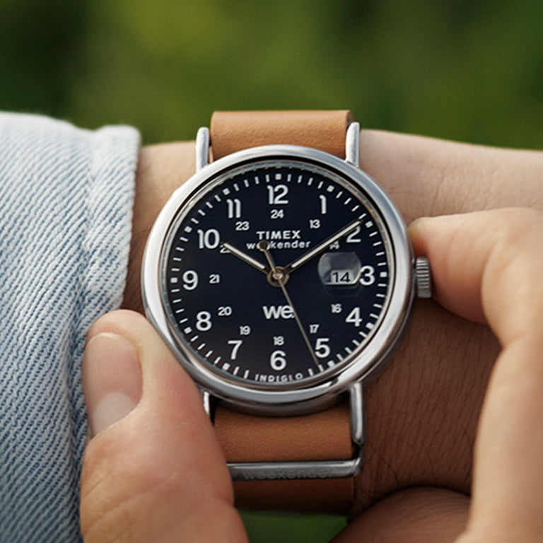 Person adjusting the crown of a Timex Weekender watch with a blue dial and brown leather strap while wearing a light blue denim shirt.