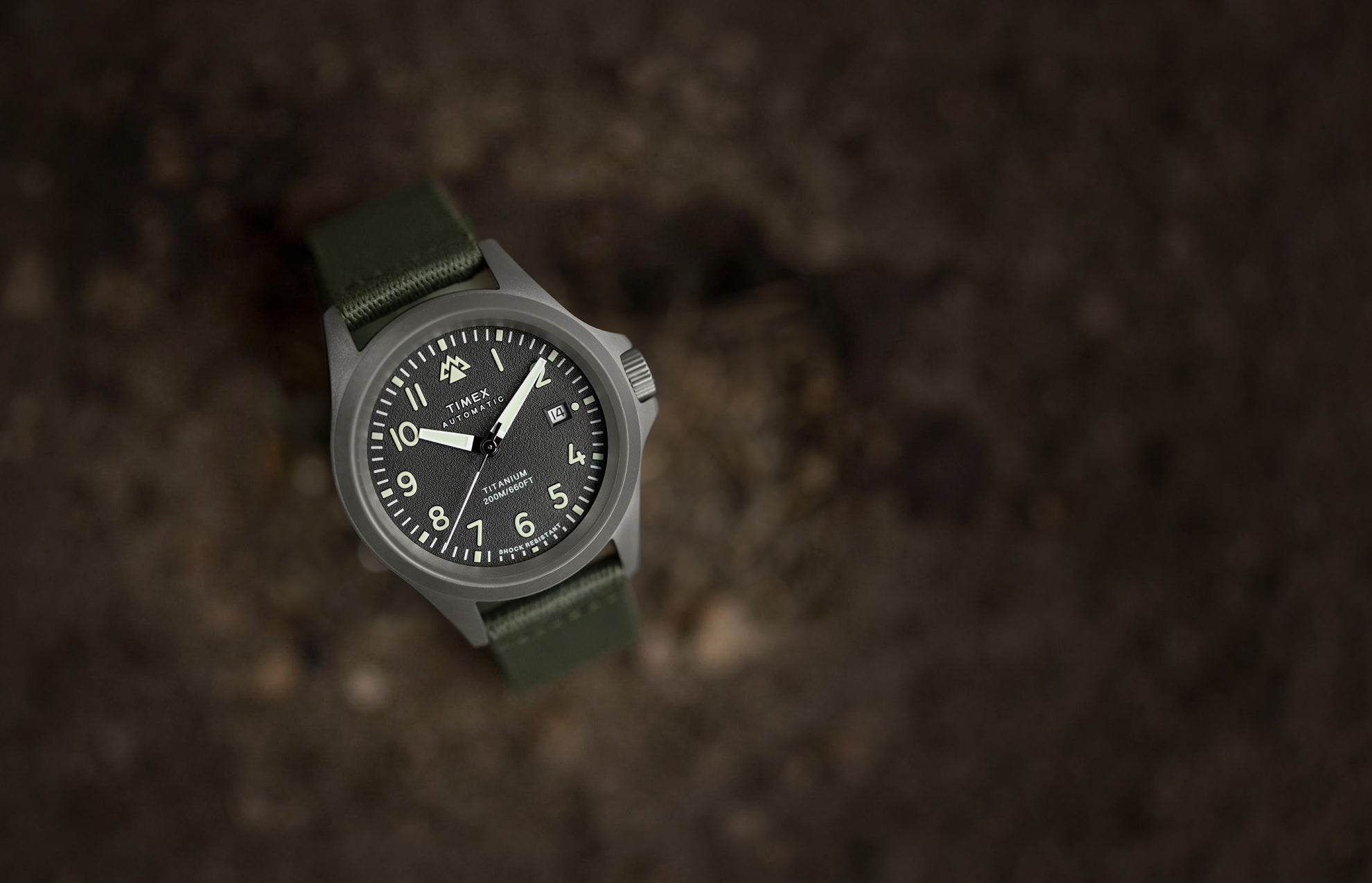 A Timex Expedition North Titanium Automatic watch with a matte titanium case, black textured dial, luminous numerals, and green fabric strap, placed on a rugged surface.