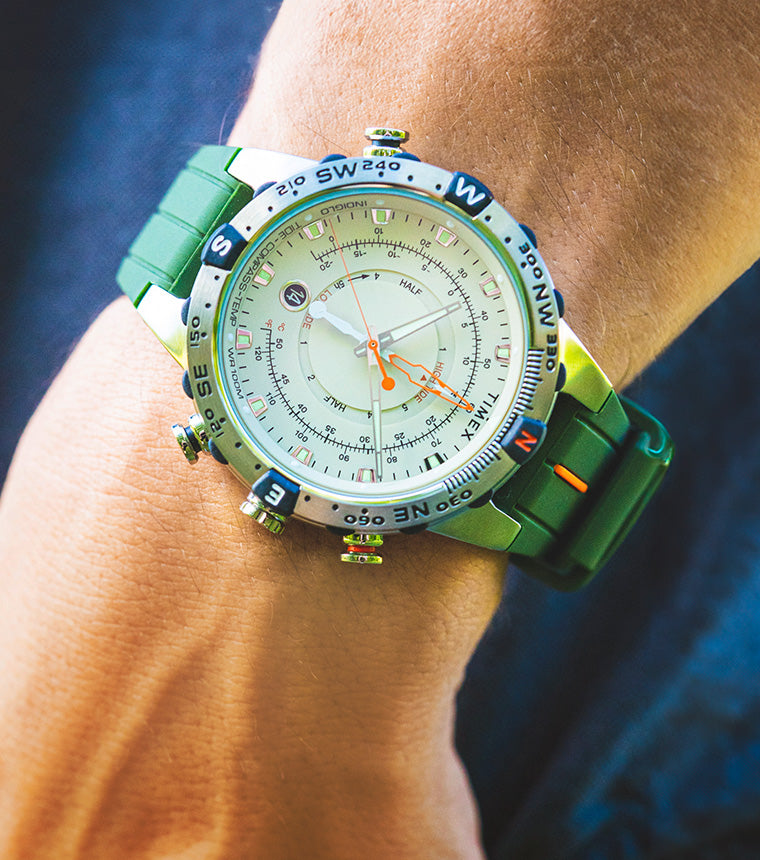 A Timex Expedition Tide-Temp-Compass watch with a cream dial, stainless-steel case, and green silicone strap worn on the wrist outdoors. Features luminous hands, orange tide indicators, and compass bezel accents.