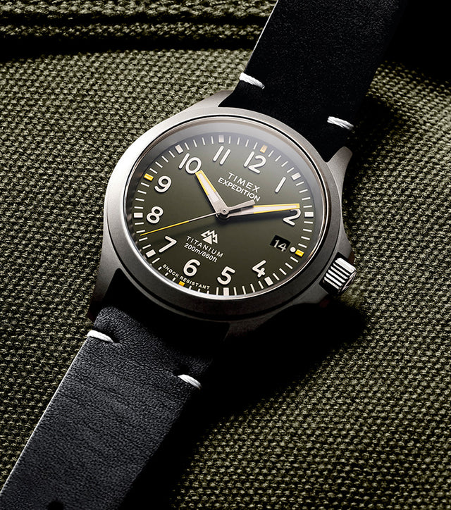 The Timex Expedition Titanium displayed flat on olive-green textured fabric, showcasing its brushed titanium case and classic field watch design.