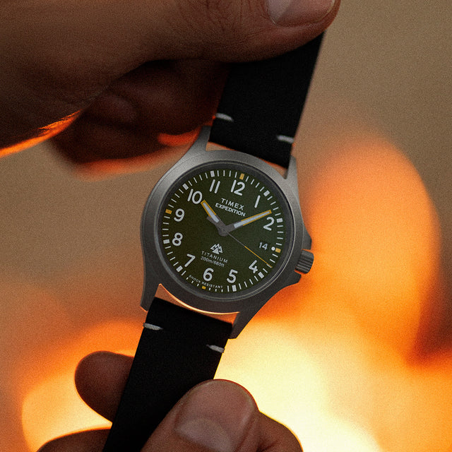 Close-up of the Timex Expedition Titanium watch being held near a fire, highlighting its luminous hands, green dial, and durable titanium build.