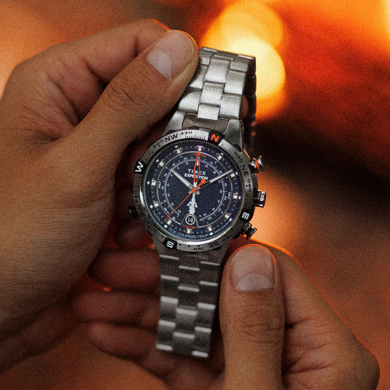 Person holding the Timex Expedition Tide-Temp-Compass in both hands, showing the stainless-steel bracelet, black multifunction dial, and compass bezel, set against a warm campfire glow.