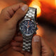 Person holding the Timex Expedition Tide-Temp-Compass in both hands, showing the stainless-steel bracelet, black multifunction dial, and compass bezel, set against a warm campfire glow.