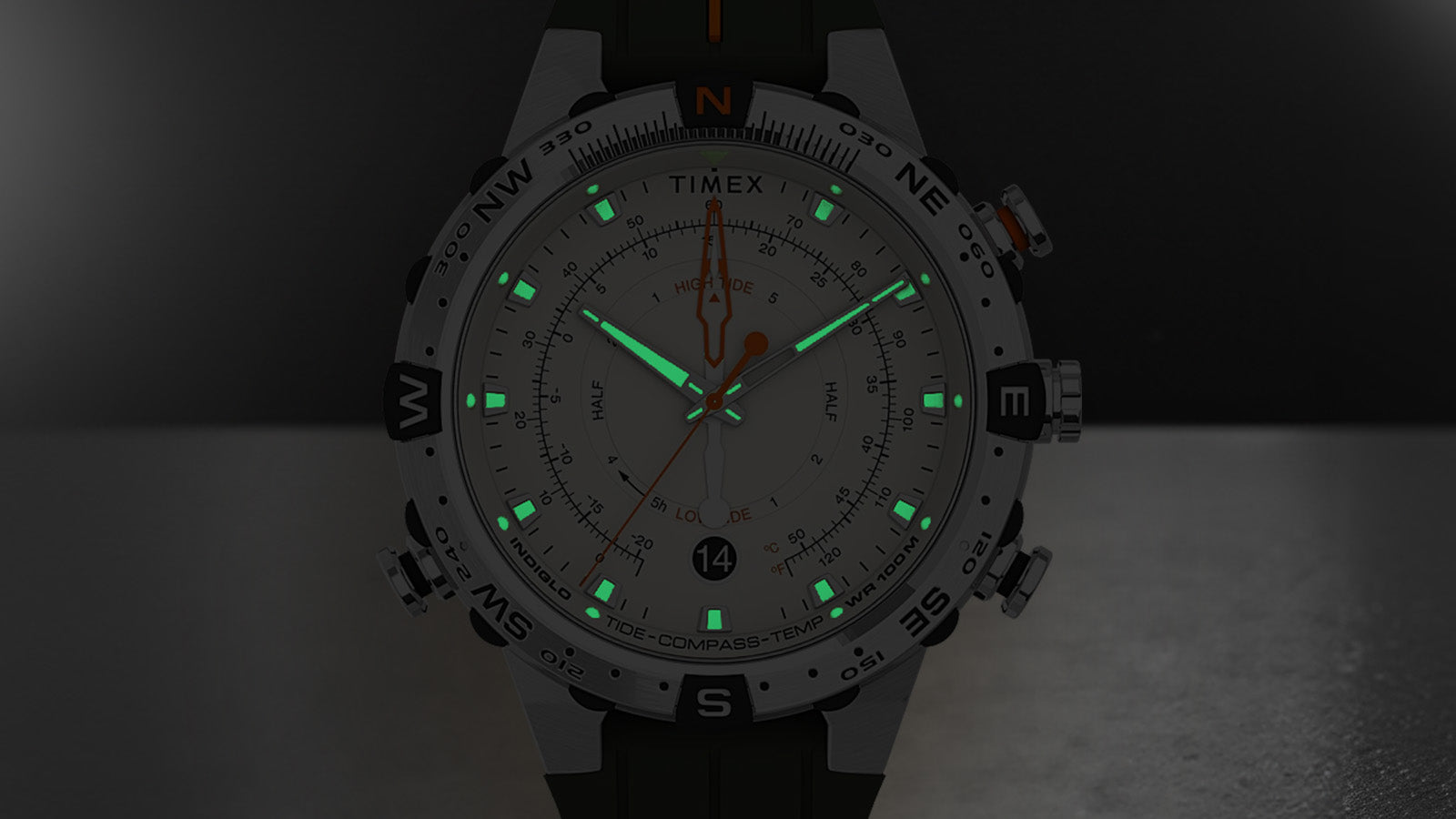 Timex Expedition Tide-Temp-Compass watch in a dark setting with Indiglo backlight and luminous green markers and hands glowing brightly against the cream dial.