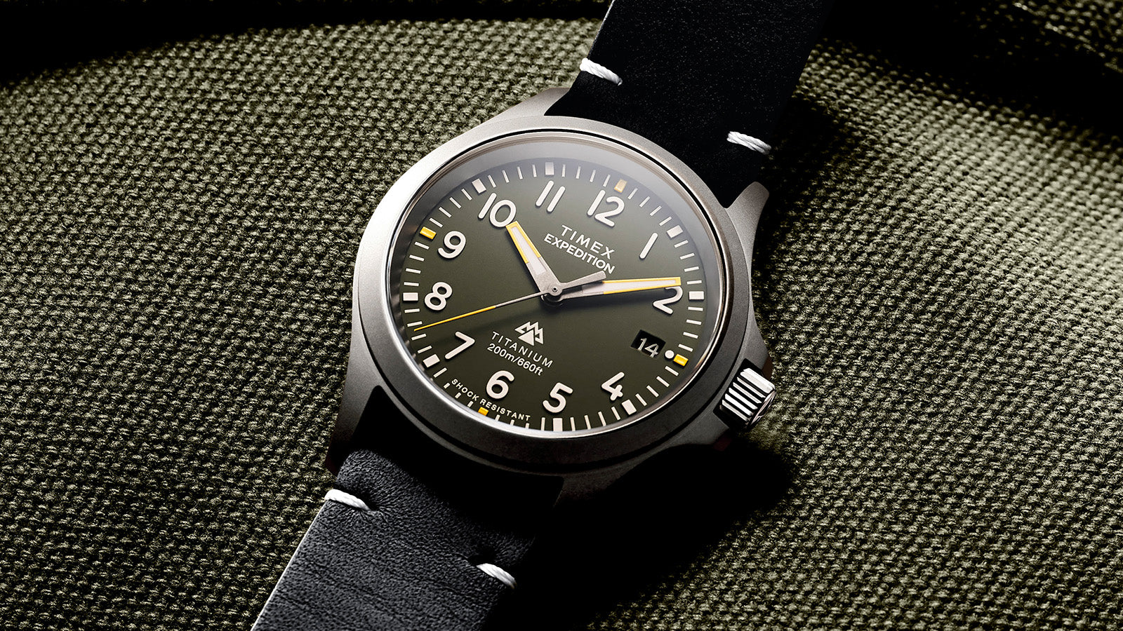 Detailed shot of the Timex Expedition Titanium on olive fabric, focusing on the green dial, white numerals, yellow second hand, and date display at 3 o’clock.