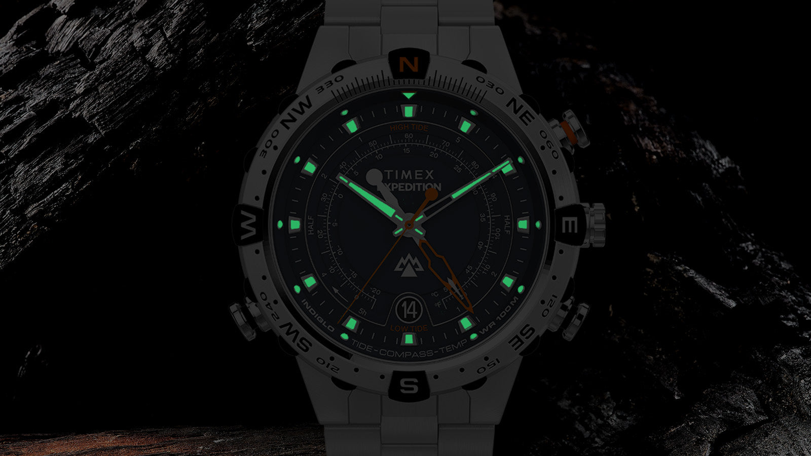 Timex Expedition Tide-Temp-Compass glowing in the dark with Indiglo backlight, green luminous markers, and compass bezel visible against a dark textured background.