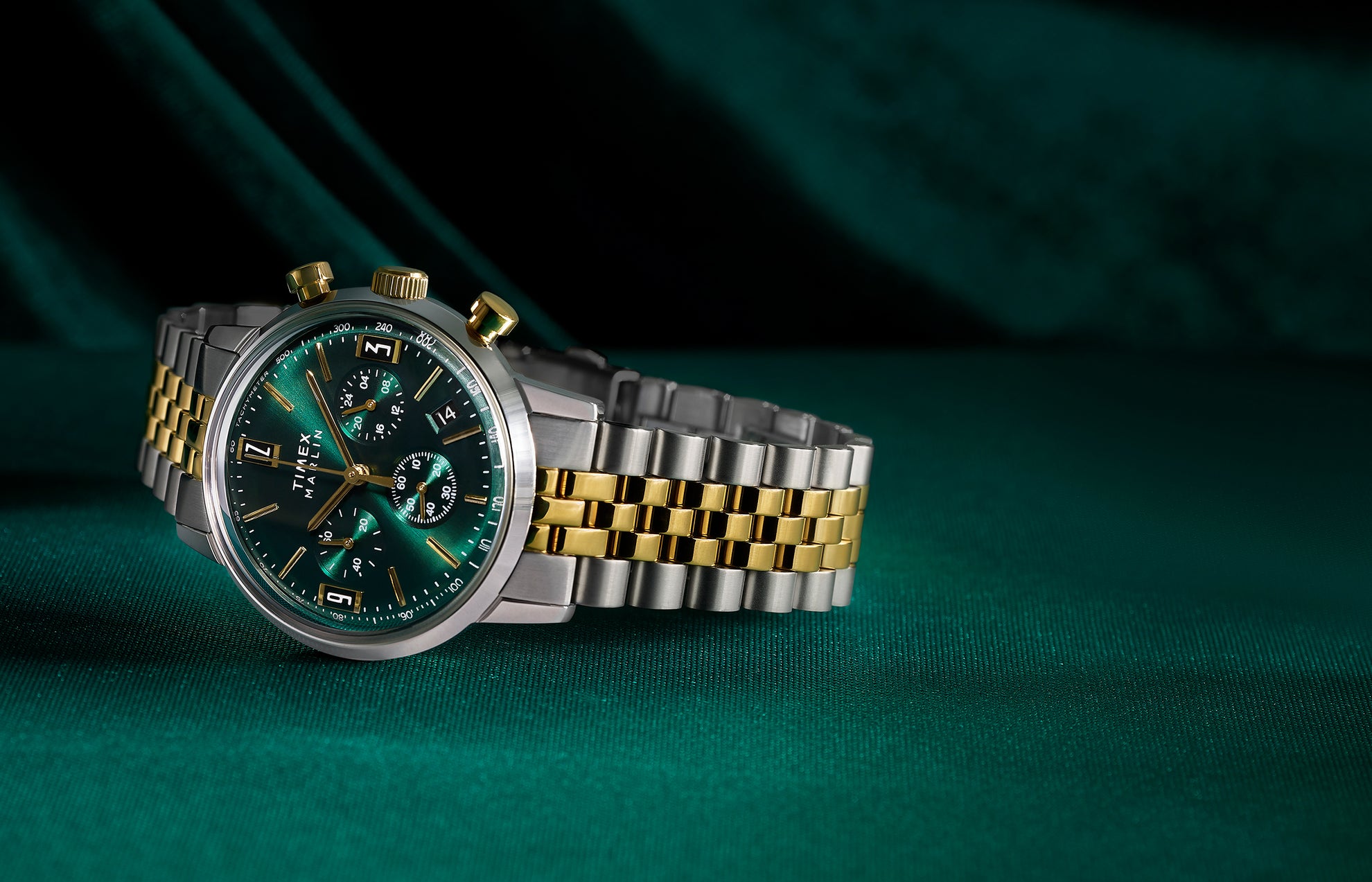 Timex Marlin Chronograph Tachymeter with emerald green dial and gold accents on a two-tone stainless-steel bracelet, displayed on rich green fabric.