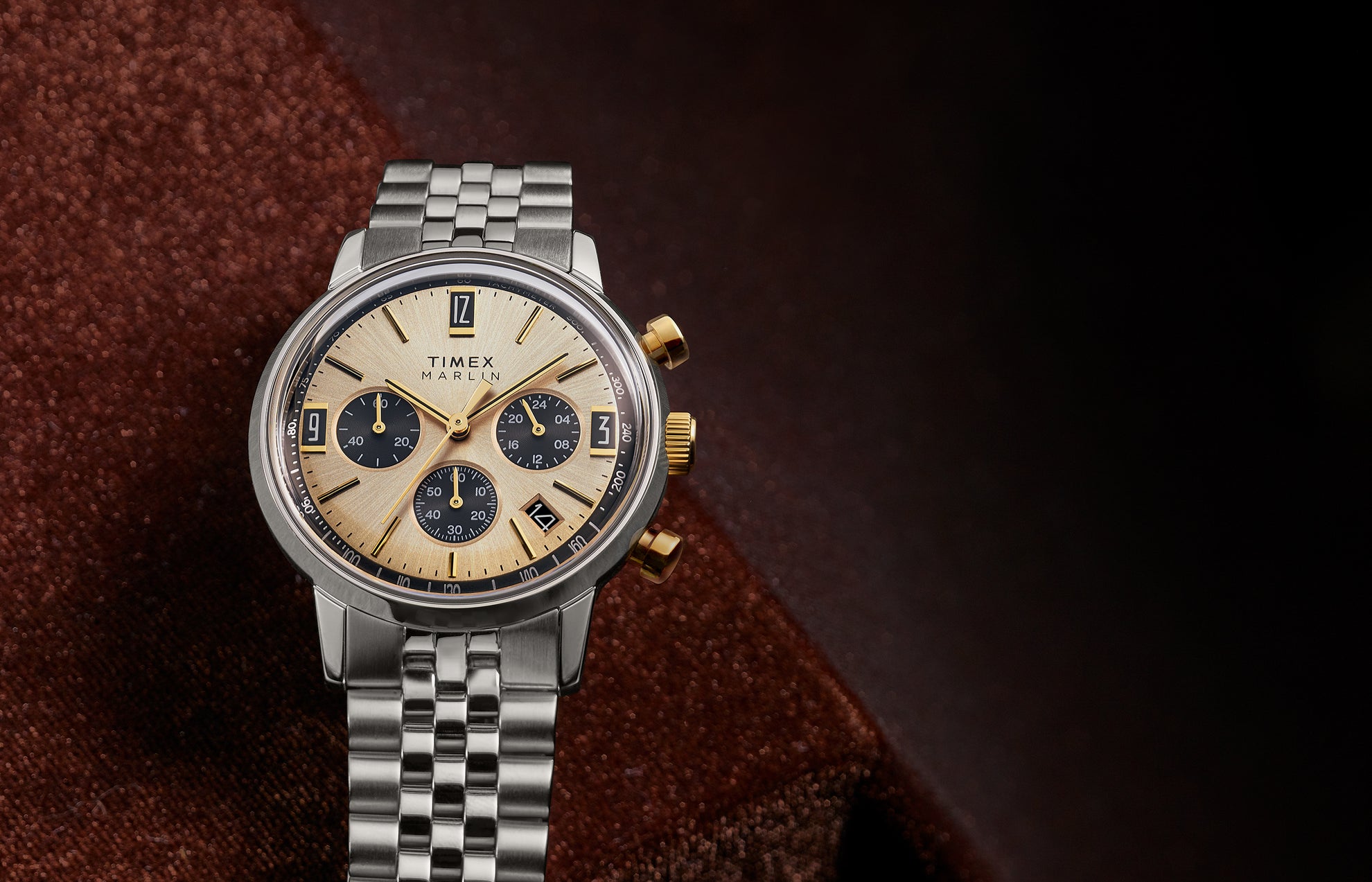 Timex Marlin Chronograph Tachymeter with champagne sunray dial and black subdials resting on rich brown fabric background.