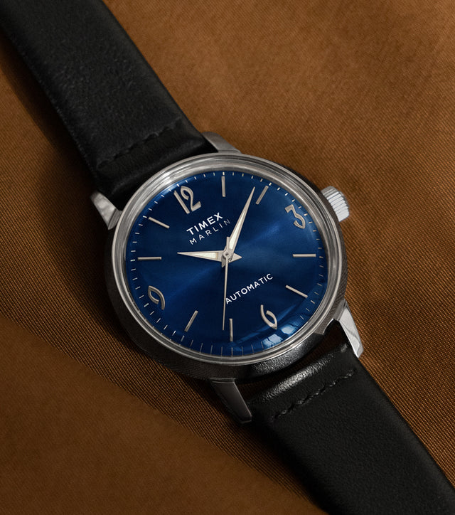 Timex Marlin Automatic watch with deep blue sunray dial and black leather strap resting on a brown fabric surface.