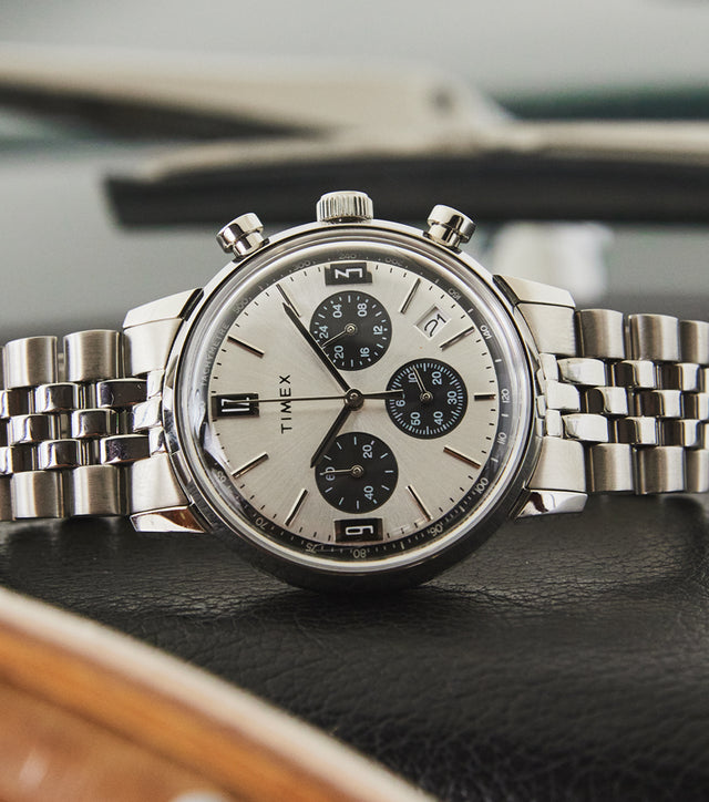 Timex Marlin Chronograph Tachymeter with silver dial, black subdials, and stainless-steel bracelet resting on black leather car dashboard.