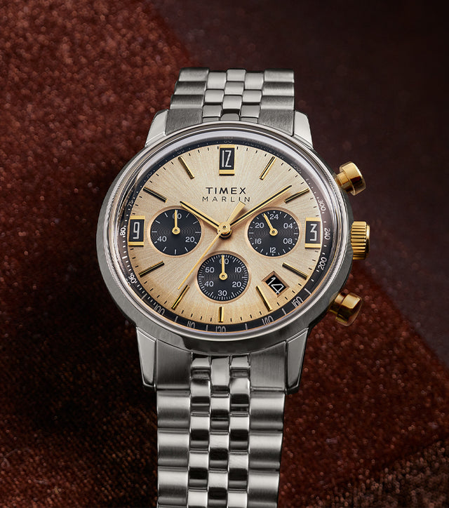 Timex Marlin Chronograph Tachymeter with champagne sunray dial and black subdials resting on rich brown fabric background.