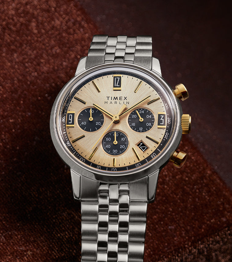 Timex Marlin Chronograph Tachymeter with champagne sunray dial and black subdials resting on rich brown fabric background.