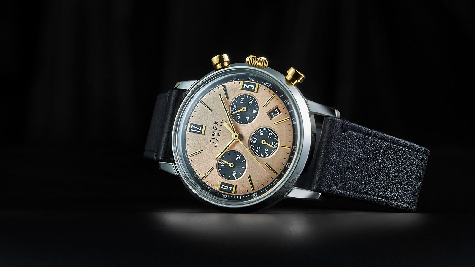 Timex Marlin Chronograph Tachymeter with champagne dial, black subdials, and gold-tone pushers displayed on black leather strap against a dark background.
