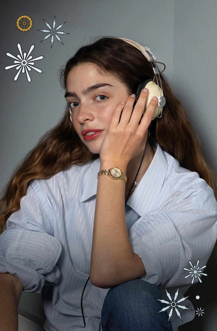 A woman wearing a light blue striped shirt and headphones smiles softly while showing her gold Timex watch, surrounded by illustrated snowflakes on a gray background.