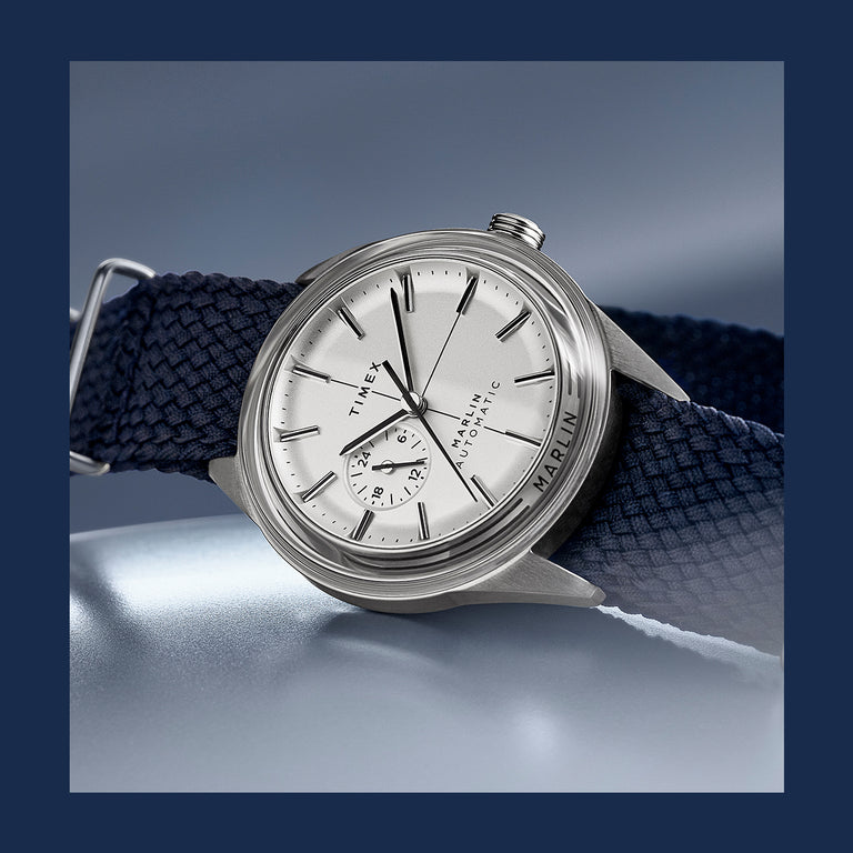 Timex Marlin Automatic watch featuring a silver dial, subdial at 9 o’clock, and navy woven strap, shown against a cool metallic background.