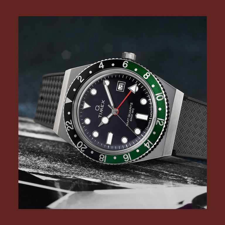 Q Timex GMT watch with a black dial, green and black bezel, red GMT hand, and textured black strap, displayed on a dark surface.