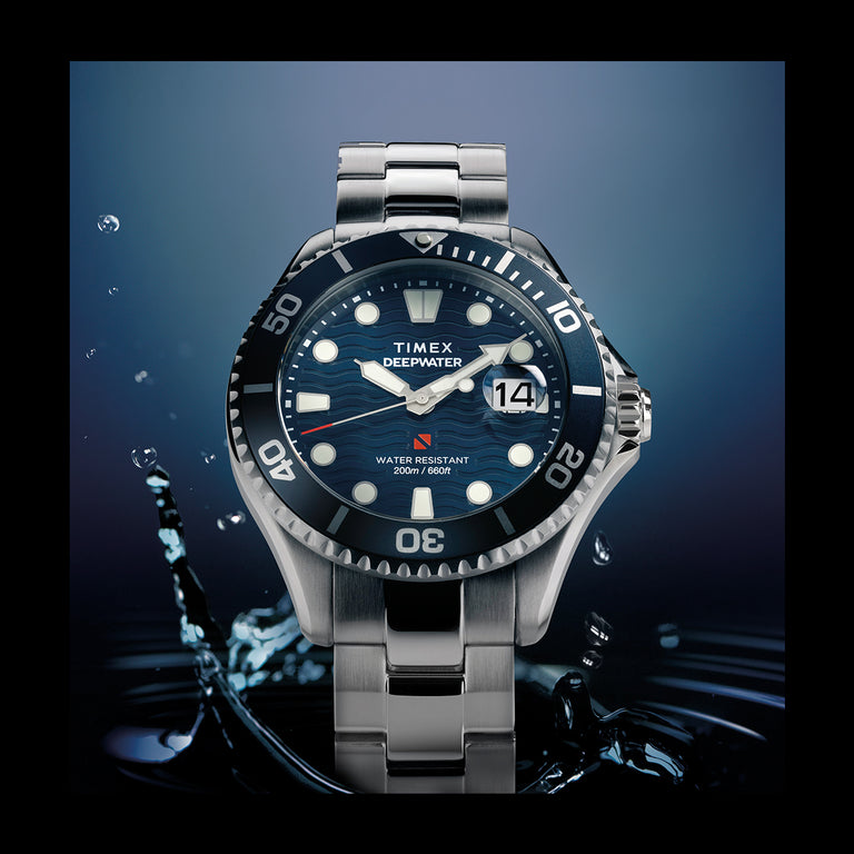 Timex Deepwater automatic dive watch with a blue wave-textured dial, luminous markers, and stainless steel bracelet surrounded by water splashes.