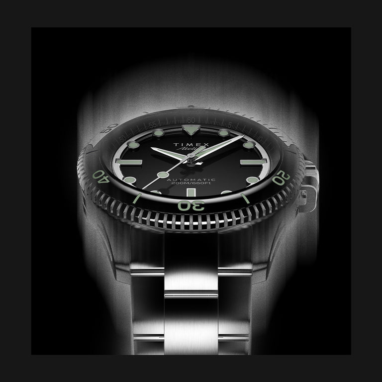 Timex Atelier watch with a black dial, luminous green markers, and stainless steel bracelet, illuminated dramatically in a monochrome lighting effect.