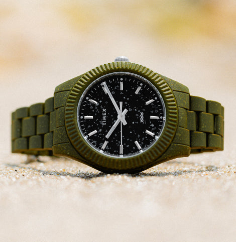 Timex eco-friendly watch featuring an olive green case and bracelet with a black speckled dial, placed on sand to highlight its sustainable design.