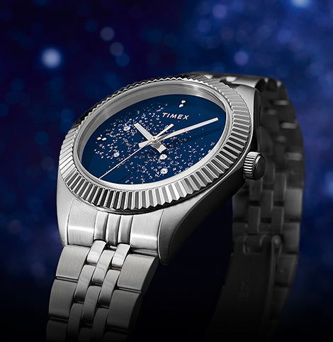 Timex watch with a blue starry dial and stainless steel bracelet, displayed at an angle against a deep blue cosmic background.