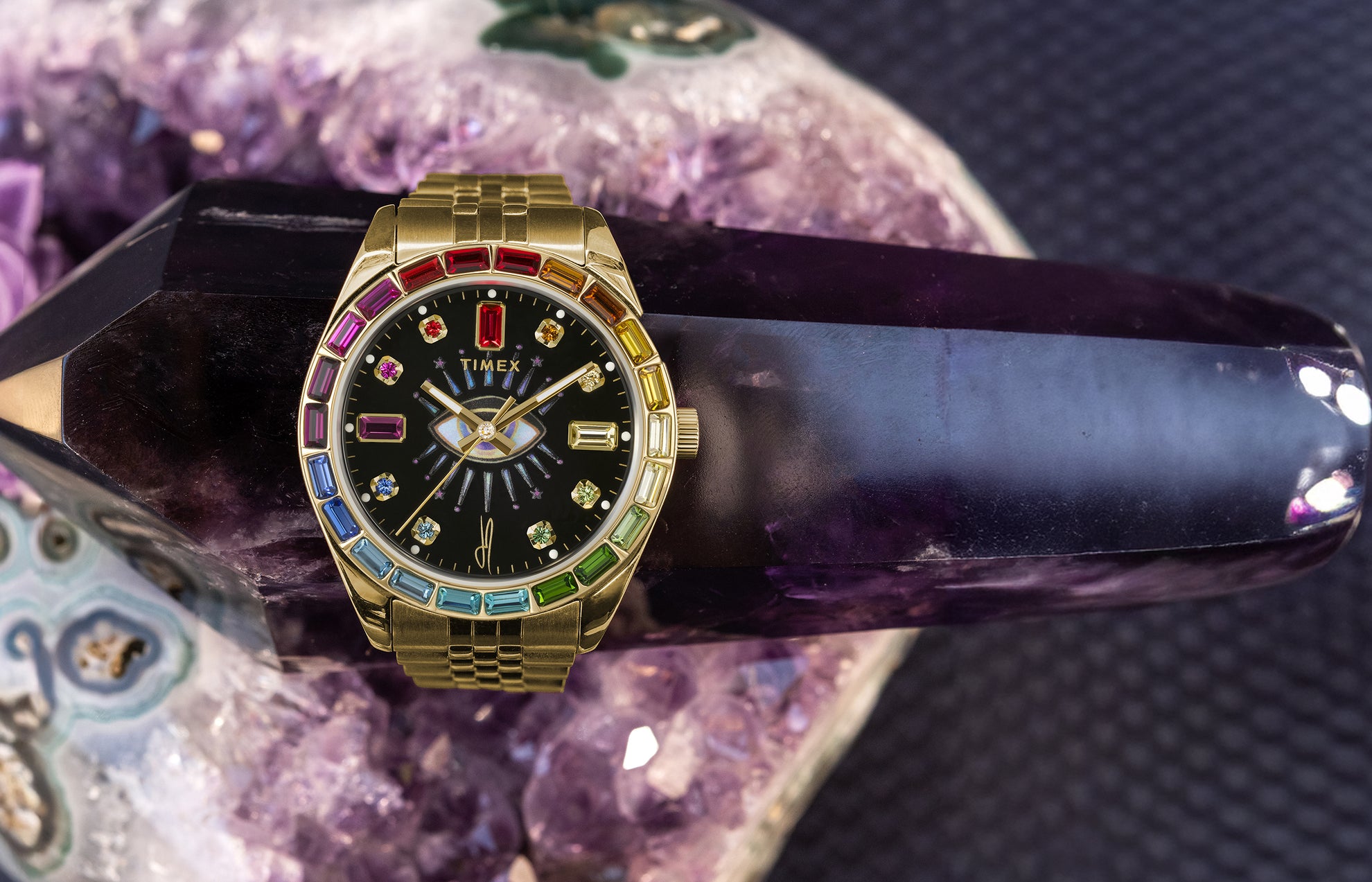 Timex x Jacquie Aiche “Cosmic Nights” gold-tone watch with a black dial adorned with rainbow-colored gemstones. The timepiece rests on an amethyst crystal, emphasizing its spiritual and luxurious aesthetic.