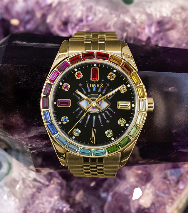 Timex x Jacquie Aiche “Cosmic Nights” gold-tone watch with a black dial adorned with rainbow-colored gemstones. The timepiece rests on an amethyst crystal, emphasizing its spiritual and luxurious aesthetic.