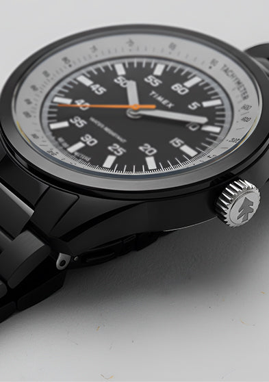 A Huckberry x Timex 1979 Black Max watch on a clean, light gray background. The black metal bracelet and case contrast with the white outer dial ring and black center dial, highlighted by an orange seconds hand.