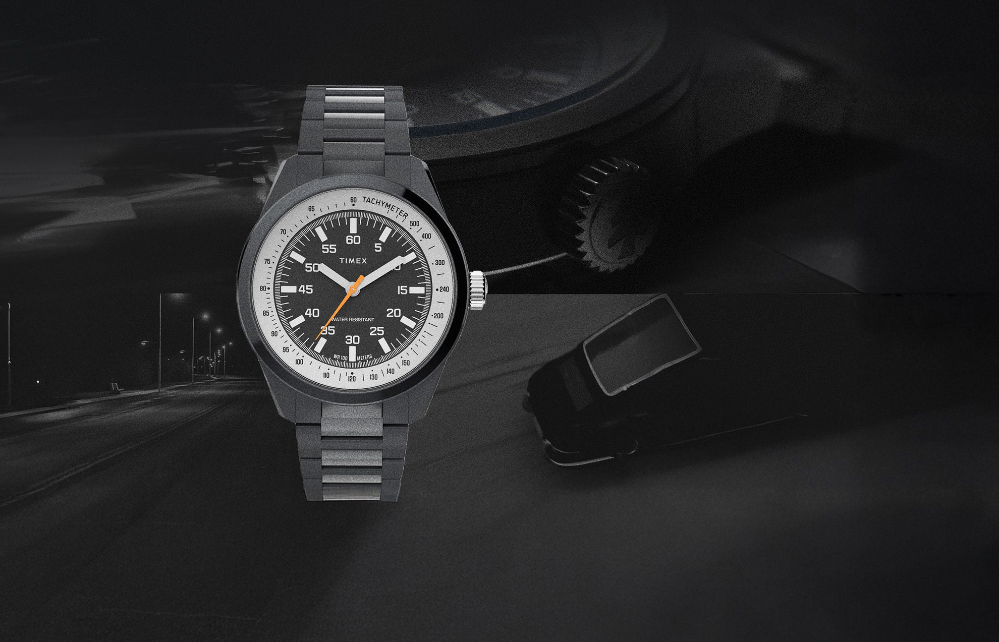 The Huckberry x Timex 1979 Black Max watch displayed against a dark, moody background featuring a night road and a faint silhouette of a car. The watch has a black case and bracelet, a black dial with white numerals, an orange seconds hand, and a tachymeter scale.