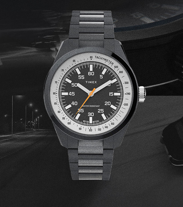 The Huckberry x Timex 1979 Black Max watch displayed against a dark, moody background featuring a night road and a faint silhouette of a car. The watch has a black case and bracelet, a black dial with white numerals, an orange seconds hand, and a tachymeter scale.