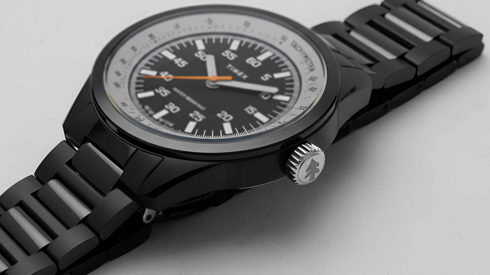 A close, side-angled shot of the Huckberry x Timex 1979 Black Max watch on a light gray background, showcasing the black bracelet links, polished case, and embossed crown logo.