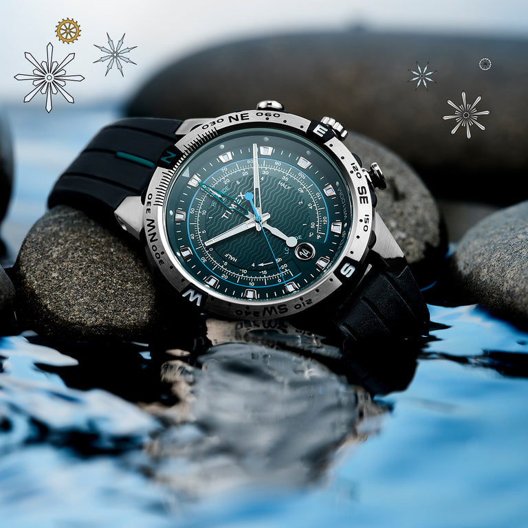 Timex Expedition watch with a black rubber strap and a detailed teal dial featuring wave patterns and multiple measurement rings. The stainless-steel bezel displays compass markings. The watch rests on smooth wet stones beside reflective water, with subtle starburst graphics around the scene.