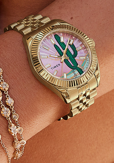 Lifestyle image of a woman wearing the Timex x Jacquie Aiche “Desert Daze” gold-tone watch with diamond and gold chain bracelets. The cactus dial glows in the sunlight, evoking bohemian luxury and desert serenity.