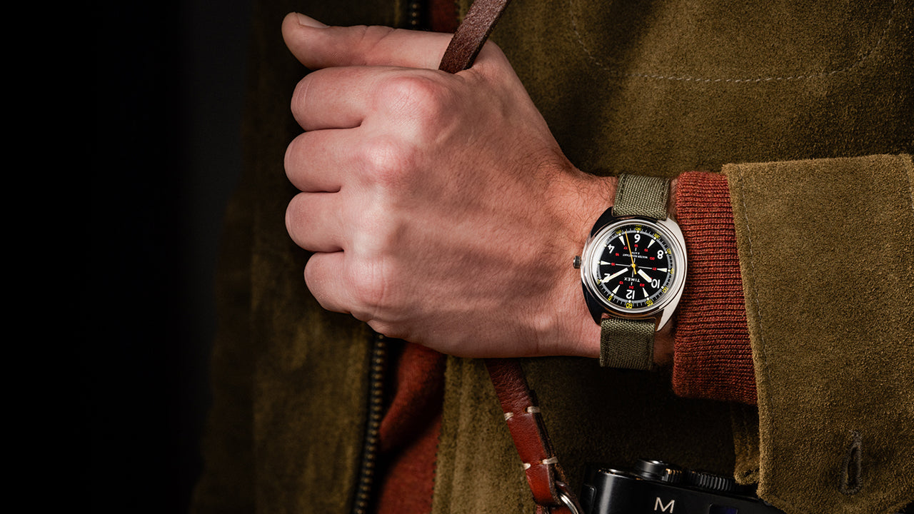 Lifestyle image of the Timex x Bespoke Post watch worn on a wrist, paired with a brown suede jacket and camera strap. The shot captures rugged sophistication and adventure-ready elegance.