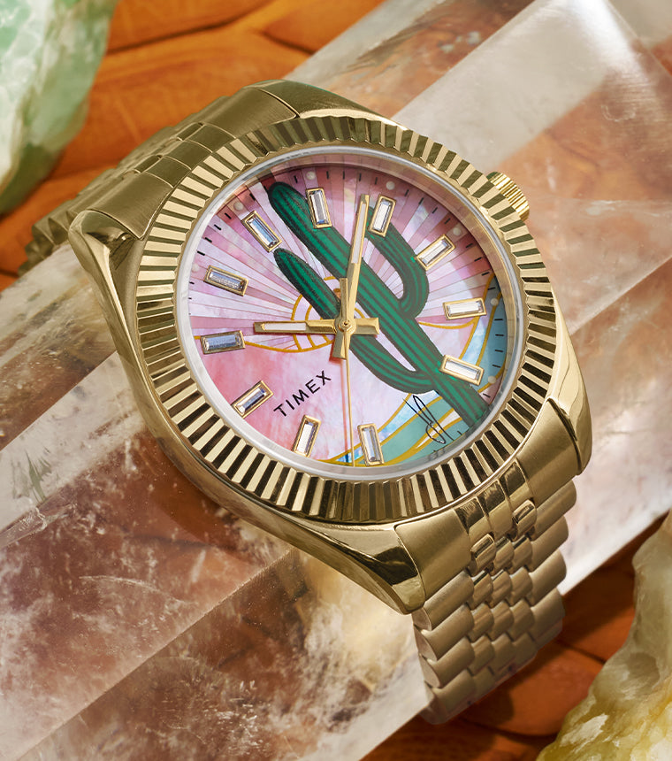 Timex x Jacquie Aiche “Desert Daze” watch with a gold-tone case and bracelet resting on large desert-toned crystals. The dial features a green cactus illustration over a pink mother-of-pearl background with gold hands and hour markers, capturing a Southwestern-inspired aesthetic.