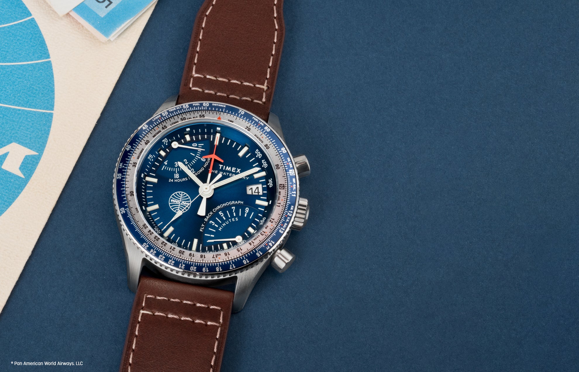 Close-up of the Timex x Pan Am® Fly-Back Chronograph watch with a blue dial, silver-tone case, and brown leather strap. The watch face features Pan Am’s globe logo, multiple sub-dials, and a rotating bezel with white and blue detailing, placed on a blue background beside vintage travel documents.