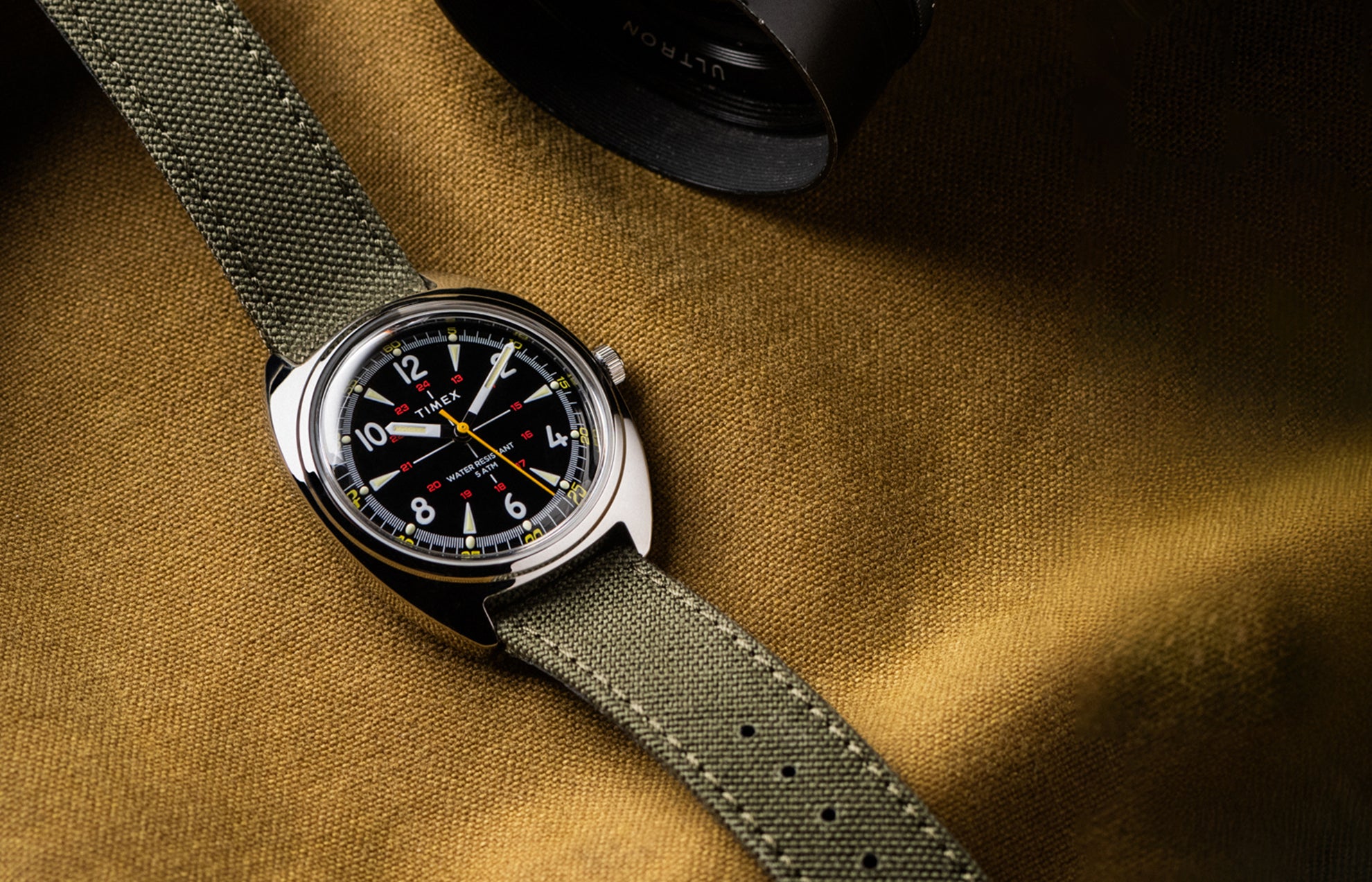 Timex x Bespoke Post watch with a black dial, luminous markers, and yellow accents, paired with an olive green fabric strap. The watch rests on a textured mustard fabric background, evoking rugged, adventure-ready style.