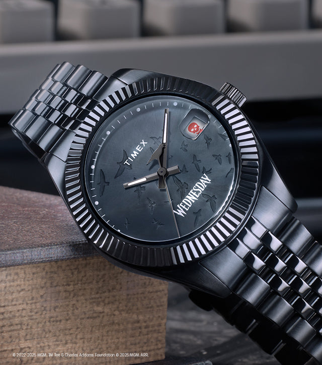 Timex x Wednesday Timex Legacy watch featuring an all-black design with a textured dial, gothic-style “Wednesday” lettering, and a skull icon date window. The watch rests on a vintage book with a dark, moody background, highlighting its mysterious aesthetic.