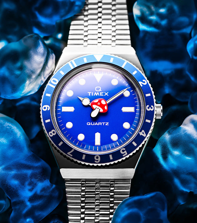 A Q Timex watch with a bright blue dial and red mushroom-shaped seconds hand, set against a background of translucent blue gummy candies. The watch has a stainless-steel bracelet and a blue bezel.
