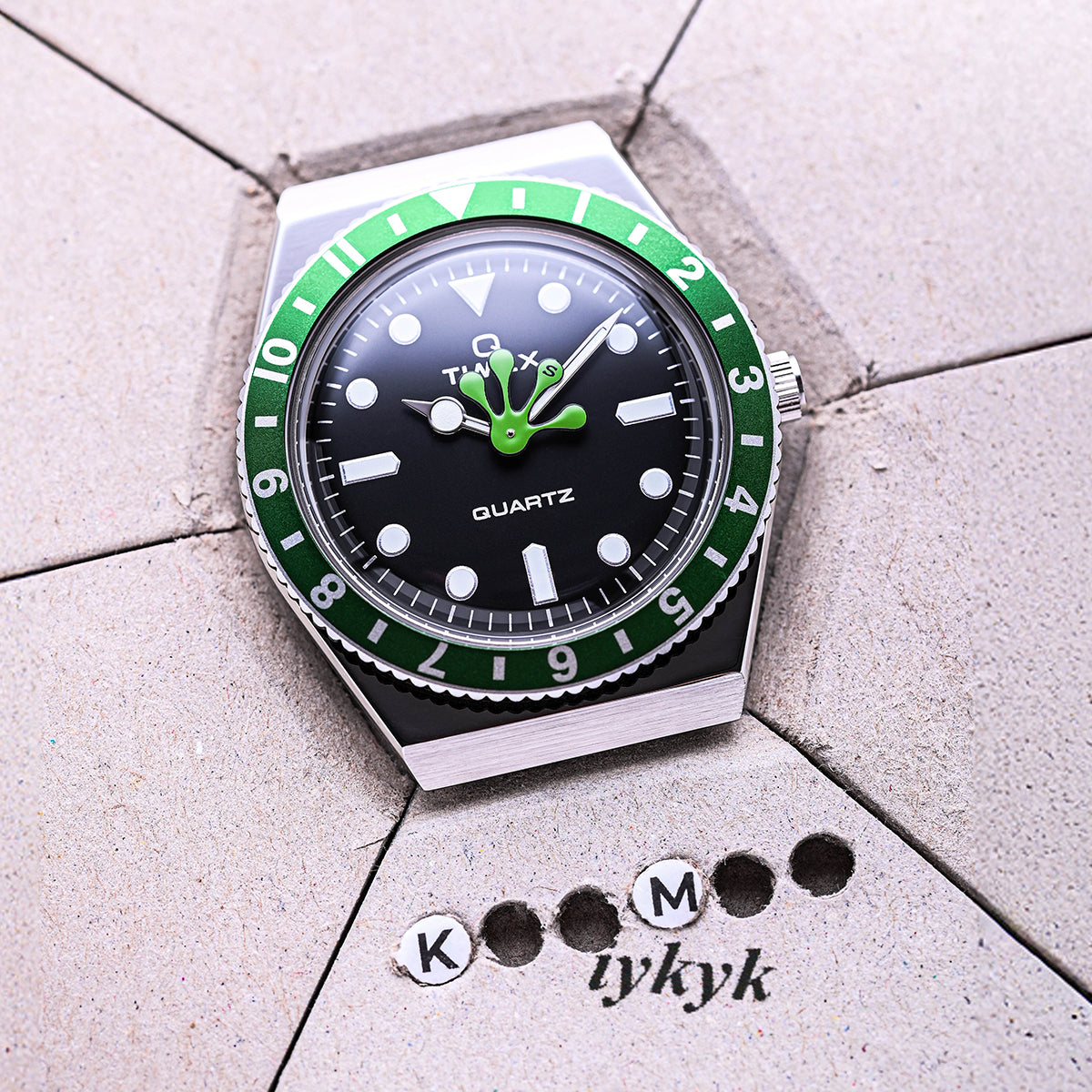 Q Timex x seconde/seconde/ watch with a green bezel and unique frog foot-shaped hour hand resting on a textured gray surface with “KM tykyk” markings. The playful collaboration merges vintage Timex design with seconde/seconde/’s witty aesthetic.