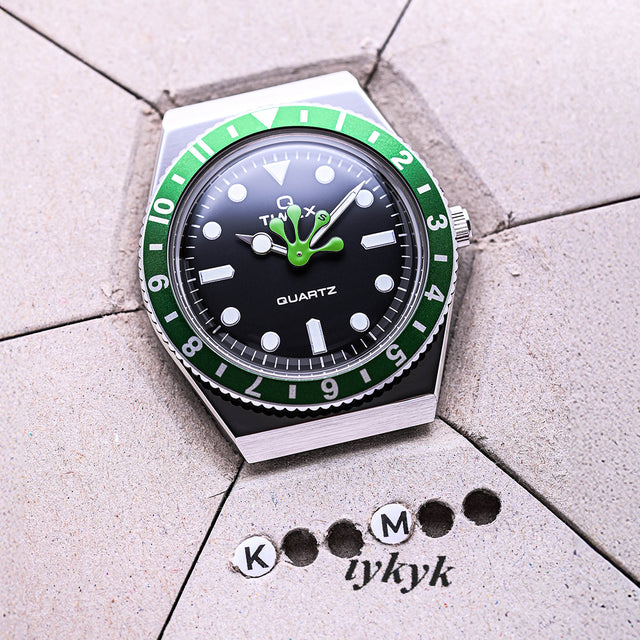 Q Timex x seconde/seconde/ watch with a green bezel and unique frog foot-shaped hour hand resting on a textured gray surface with “KM tykyk” markings. The playful collaboration merges vintage Timex design with seconde/seconde/’s witty aesthetic.
