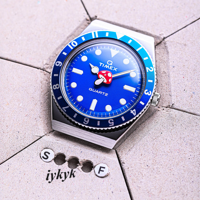 A close-up of the Q Timex blue-dial watch with a red mushroom seconds hand resting on a textured gray surface with the text “iykyk” and punched letters “S” and “F.”
