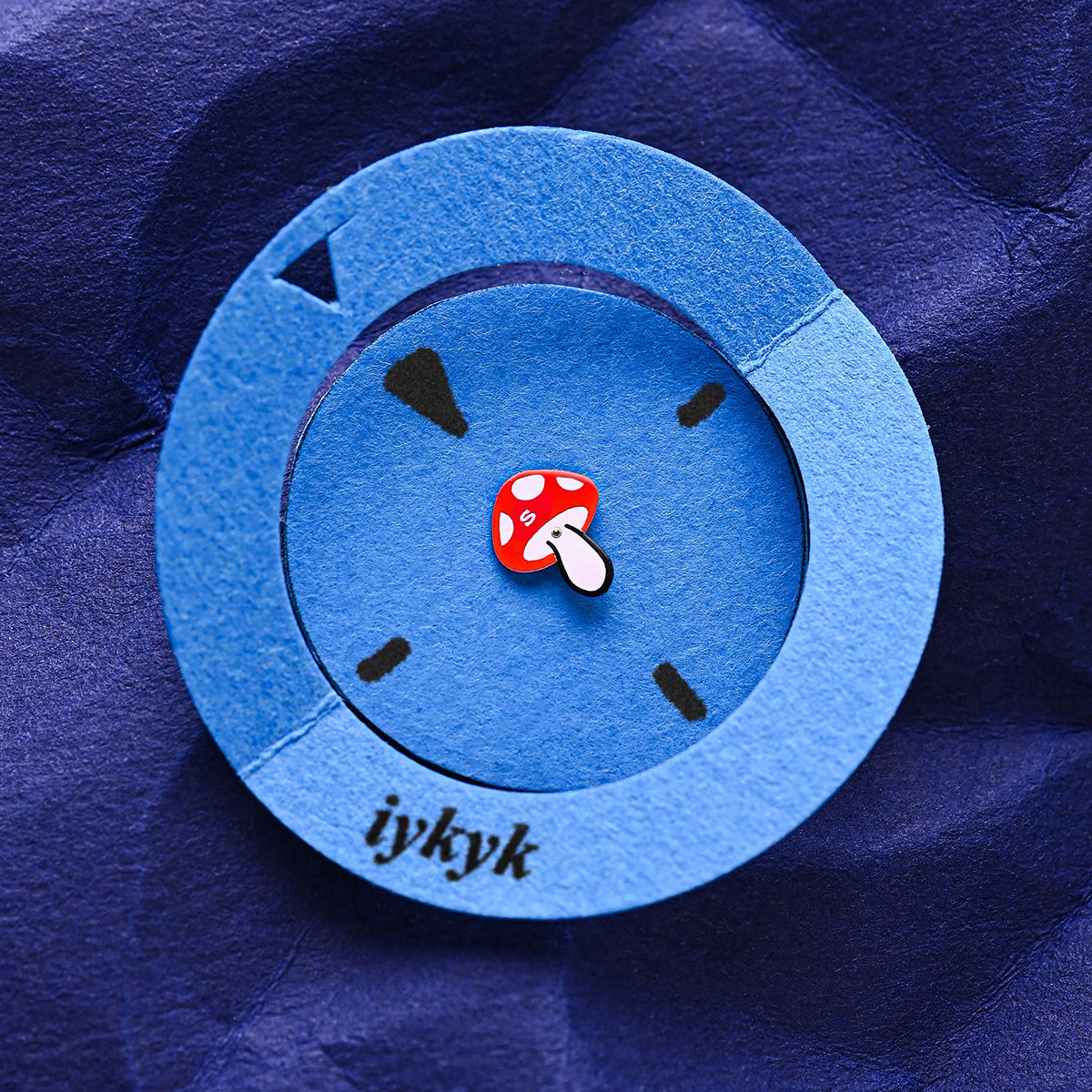 A paper art version of the Q Timex watch face, with a blue circular cutout and a red mushroom seconds hand on a deep blue textured background, featuring the word “iykyk.”