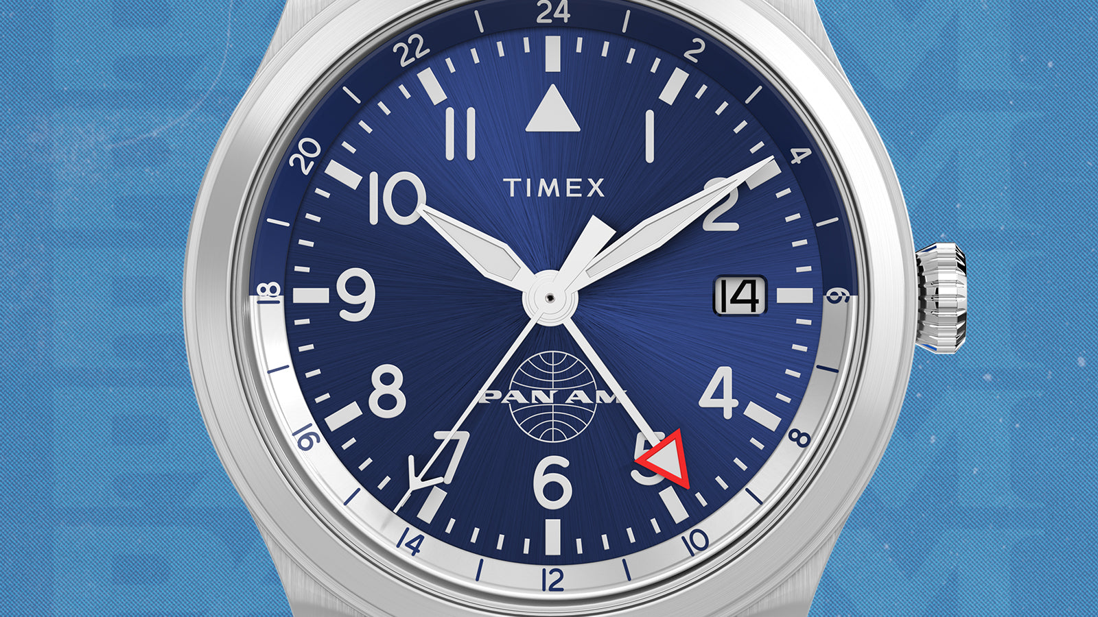 Macro shot of the Timex x Pan Am® Waterbury GMT watch dial. The deep blue sunray finish features bold white numerals, a red GMT hand, date window at 3 o’clock, and the Pan Am globe logo at 6 o’clock — blending heritage design with global travel functionality.