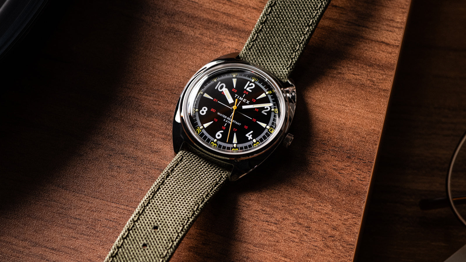 Detailed angled shot of the Timex x Bespoke Post watch on a wooden surface, emphasizing the domed crystal, smooth bezel, and fine texture of the green strap. A classic field watch design blending modern durability with retro charm.